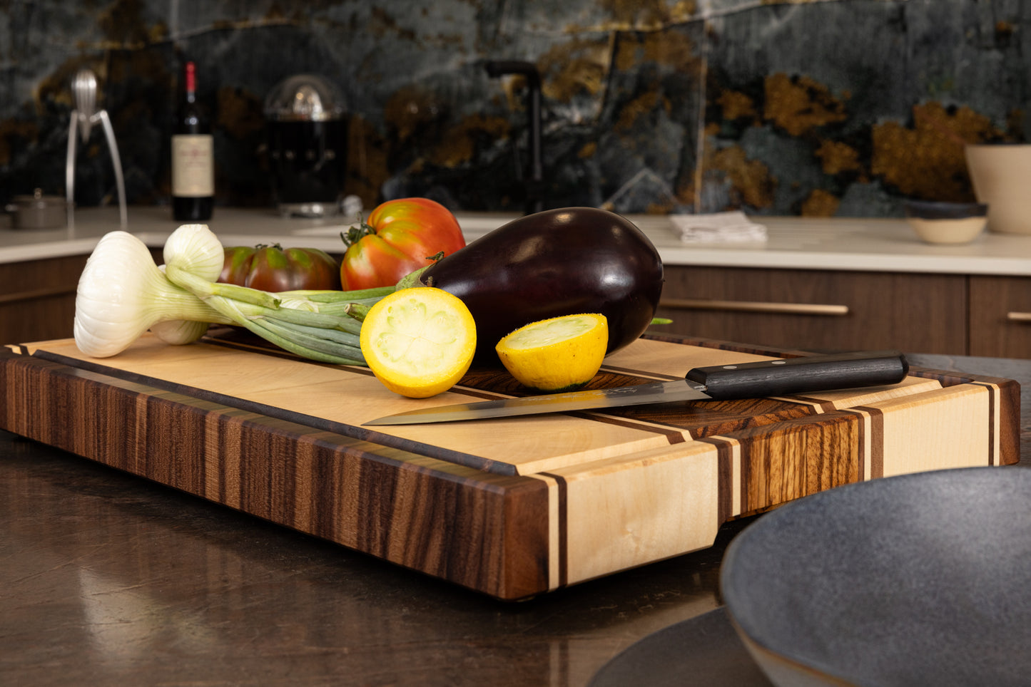Planche à découper artisanale en bois haut de gamme, modèle Couperin en zebrano, érable et noyer américain, posée sur un plan de travail de cuisine avec légumes frais et couteau de chef