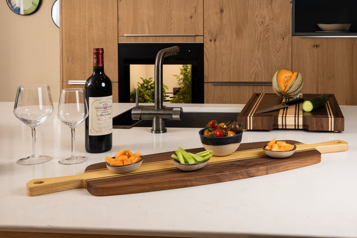 Planche apéro artisanale en n oyer et érable massif avec double poignée, présentation de légumes frais et melon, accompagnée de vin rouge dans une cuisine moderne