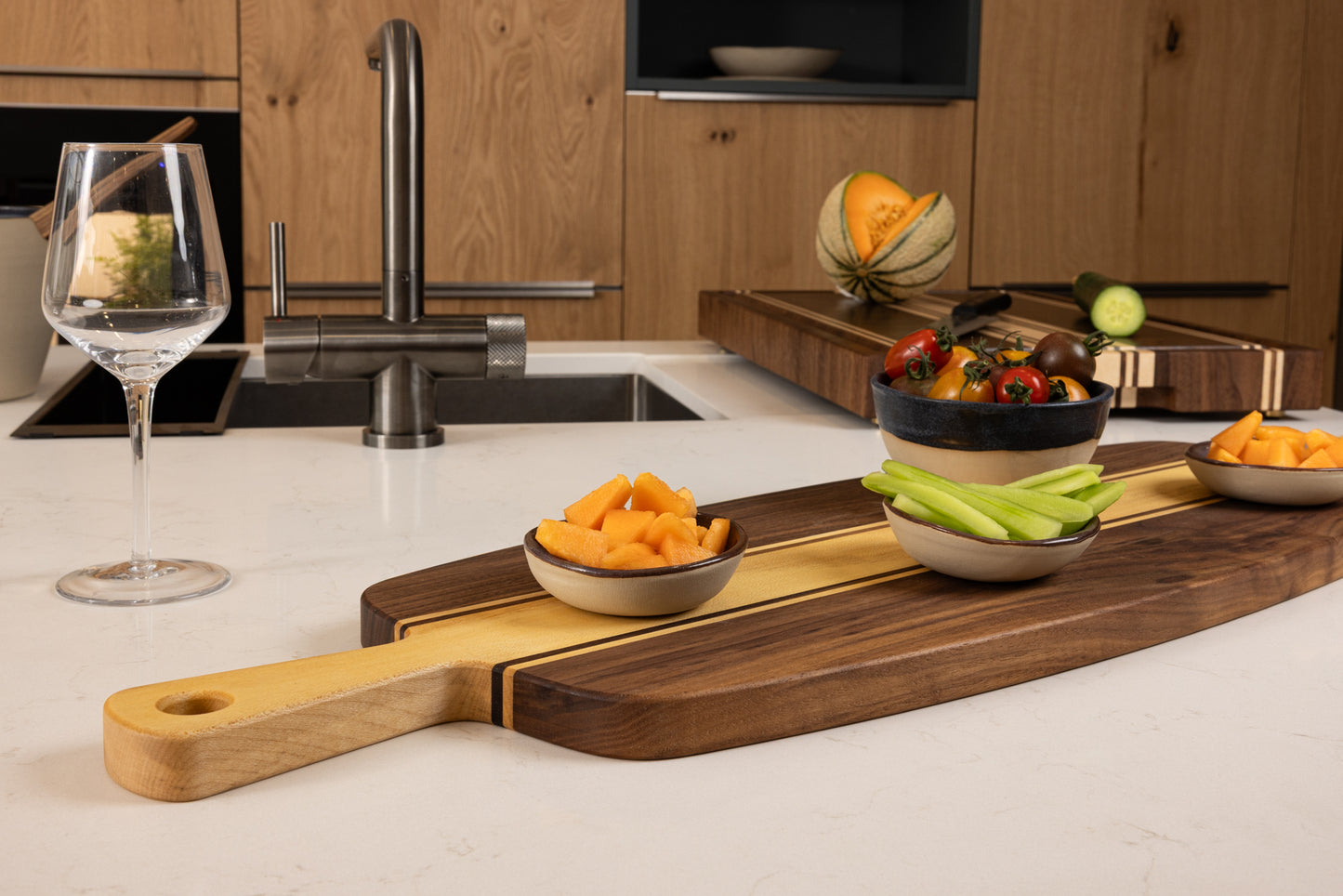 Planche apéro artisanale en noyer et érable massif avec double poignée, présentée en situation sur un plan de travail, garnie de légumes frais et de fruits à partager.