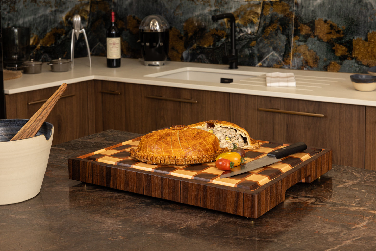 Planche en bois de bout artisanale, en poirier, noyer et érable en situation sur un plan de travail de cuisine, avec une tourte et couteau de cuisine