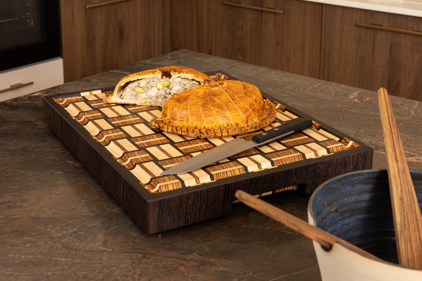 Planche à découper en bois de bout artisanale en zebrano, noyer et érable, présentée dans une cuisine avec une tourte rustique et un couteau de chef