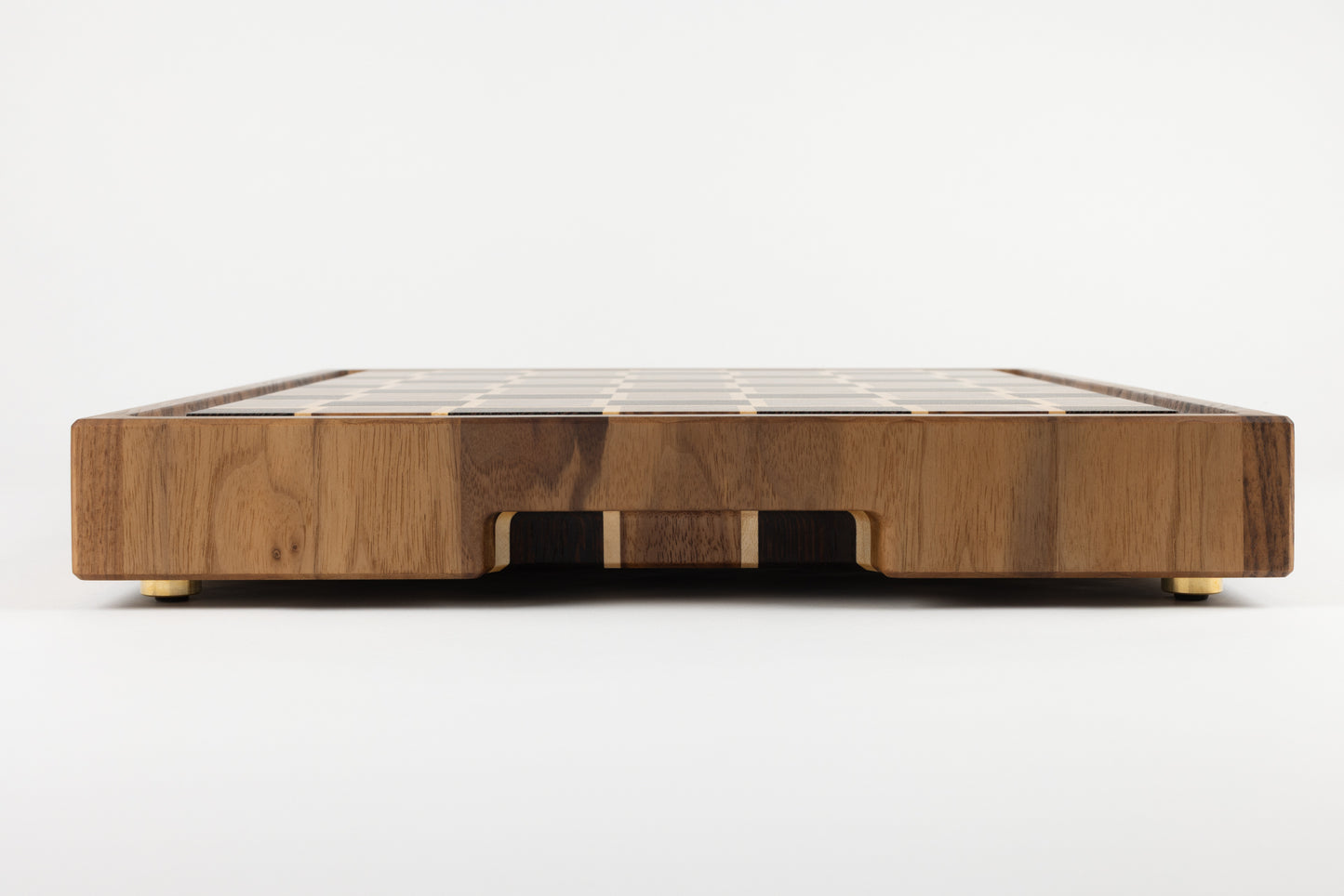 Planche à découper artisanale de luxe en bois de bout, vue de tranche sur la bordure en noyer, détail de la poignée fraisée et pieds en laiton massif fabriqués en France. »