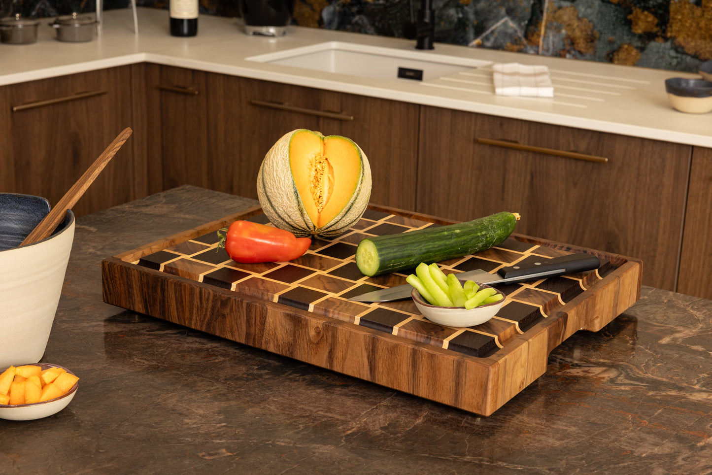 Planche à découper en bois de bout modèle Berlioz, en wengé, noyer et érable, posée sur un plan de travail en granit. Mise en scène avec légumes frais et couteau de chef, pièce artisanale française en bois haut de gamme