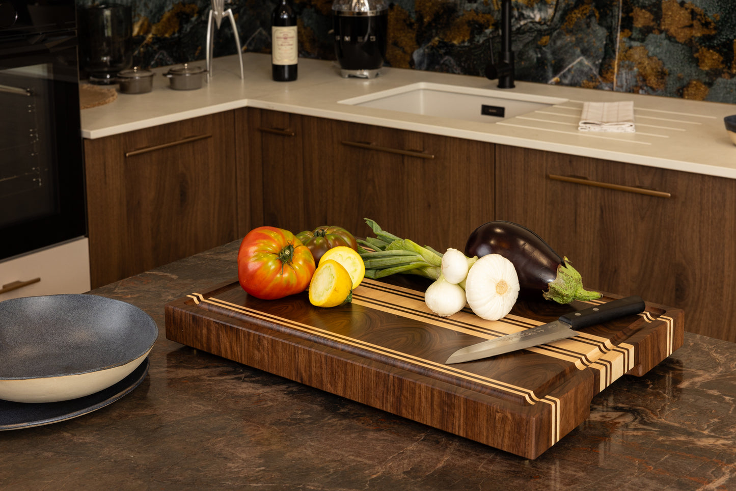 Planche à découper Debussy en noyer américain et érable, design indémodable et élégant, mise en situation dans une cuisine haut de gamme avec légumes frais et couteau de cuisine.