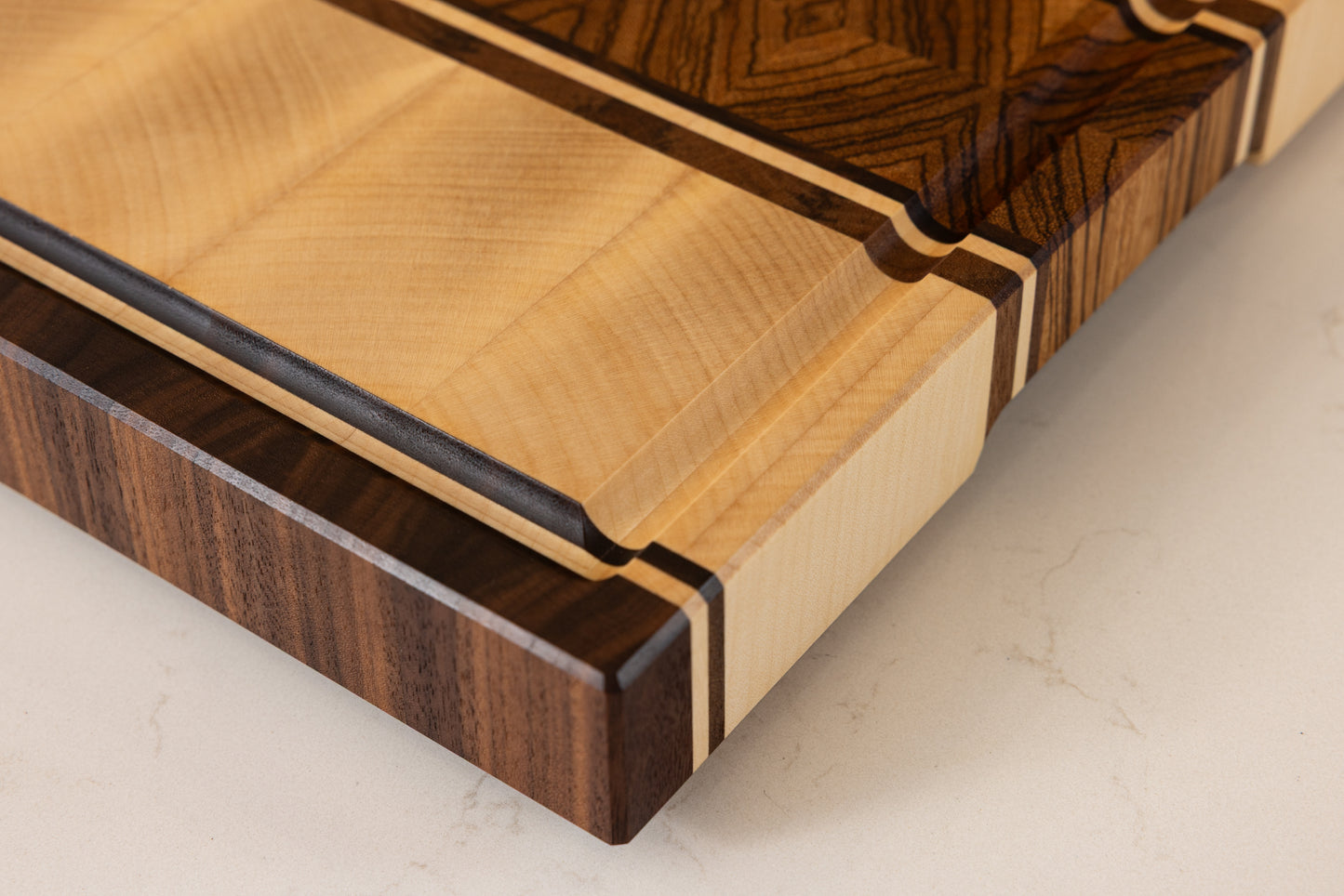 Détail d’une planche à découper artisanale en bois massif, modèle Couperin, montrant le coin avec rigole à jus sculptée, en zebrano, érable et noyer américain, posée sur un plan de travail de cuisine