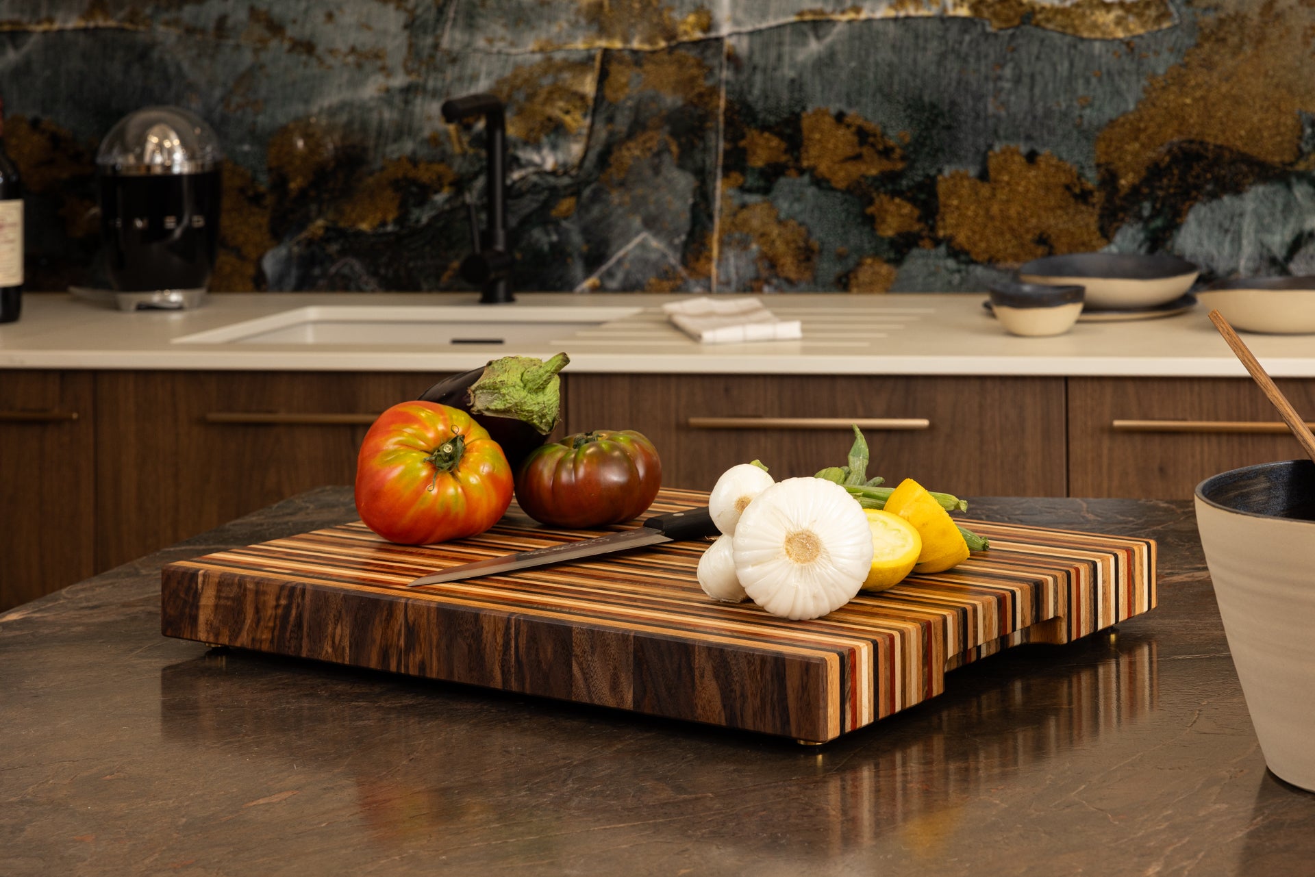 Planche à découper modèle Signature réalisée par un artisan d’art, associant 8 essences de bois nobles, mise en scène dans une cuisine haut de gamme avec légumes frais et couteau, fabrication Made in France.