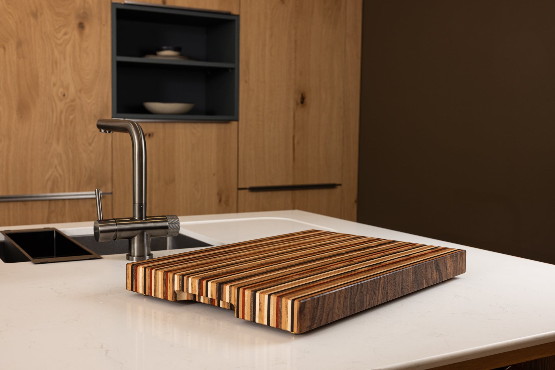 Planche à découper de luxe modèle Signature en 8 essences de bois nobles, vue de côté avec la poignée fraisée et arrière-plan d’une cuisine moderne haut de gamme en chêne.