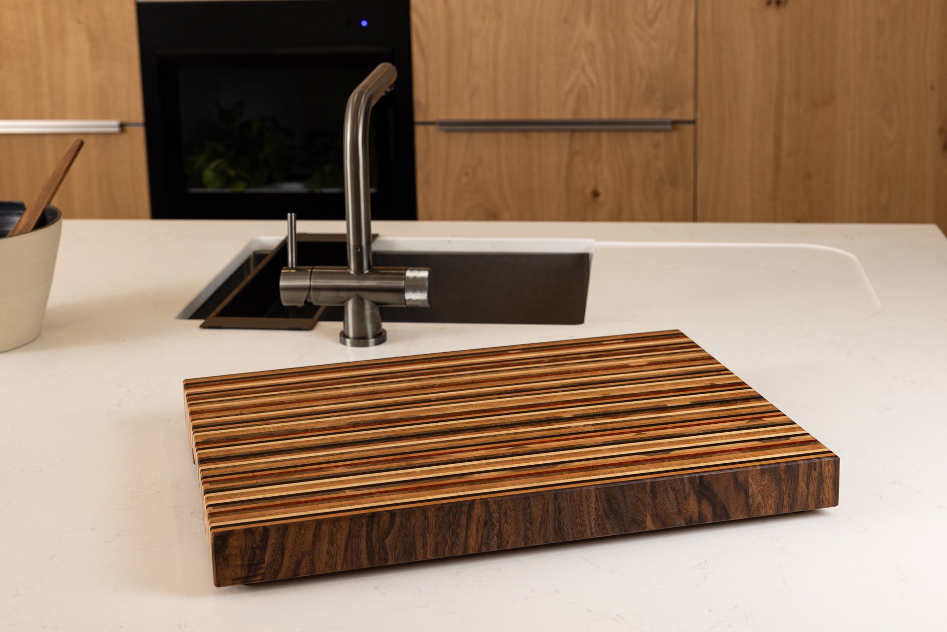 Planche à découper artisanale premium modèle Signature en 8 essences de bois nobles, posée sur un plan de travail dans une cuisine moderne élégante en chêne, création Made in France d’un artisan d’art.