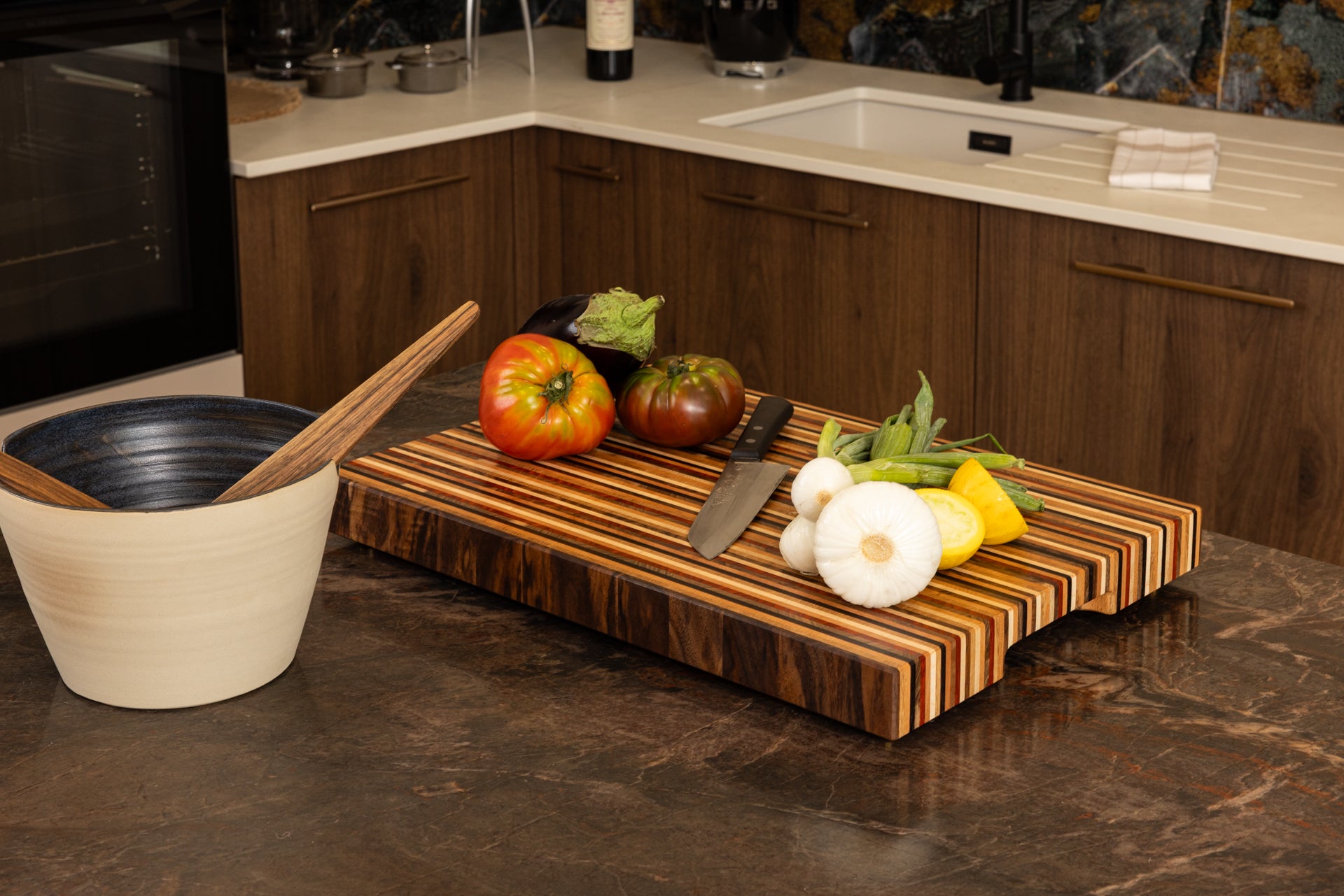 Planche à découper de luxe modèle Signature, design graphique en 8 essences de bois nobles, en situation dans une cuisine gastronomique haut de gamme avec couteau de chef et légumes frais.