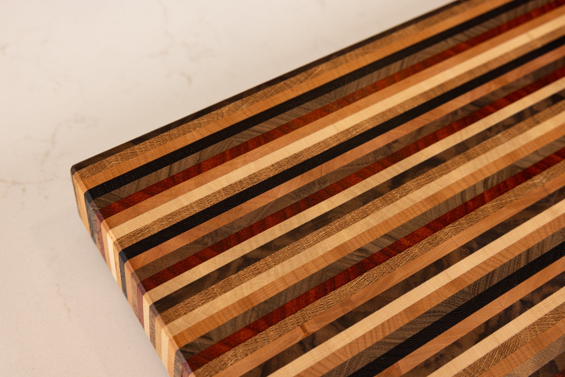 Gros plan sur le coin de la planche à découper Signature, design graphique en bandes fines de bois de bout, associant 8 essences nobles : hêtre, chêne, noyer français et américain, wengé, padouk, érable et poirier, création d’un artisan d’art Made in France. »