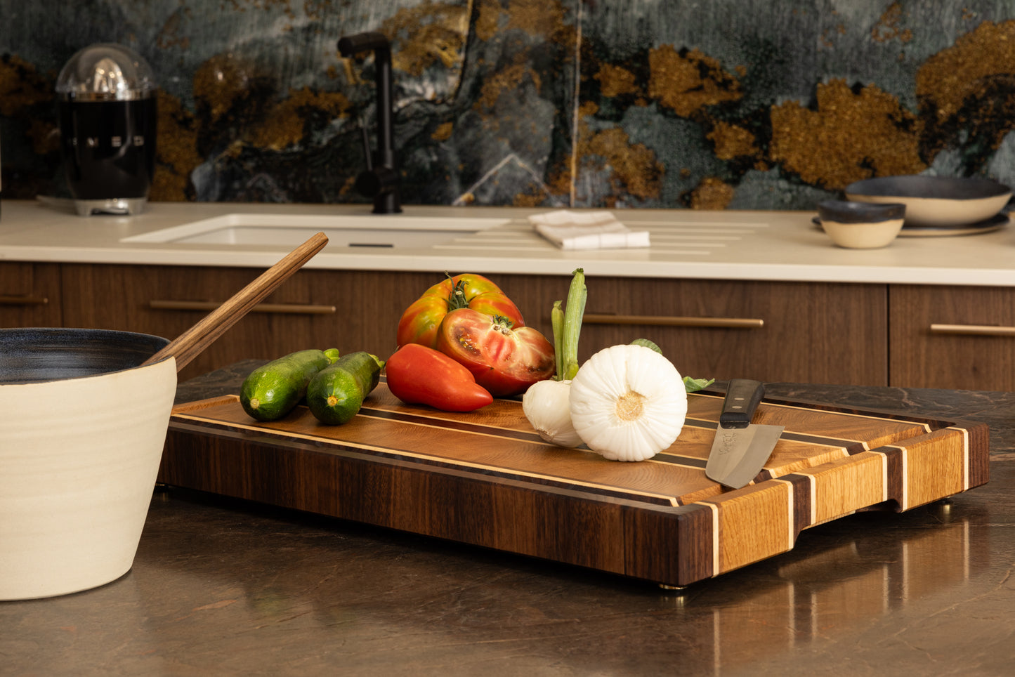 Vue en perspective de la planche à découper Ravel en chêne, noyer et érable, sur un plan de travail en granit. Présentée avec des légumes et un couteau de chef, fabrication artisanale en France