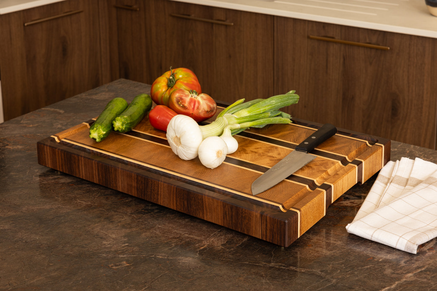 Planche à découper en bois de bout modèle Ravel, en chêne, noyer et érable, posée sur un plan de travail en granit avec légumes frais et couteau de chef. Fabrication artisanale française en bois nobles