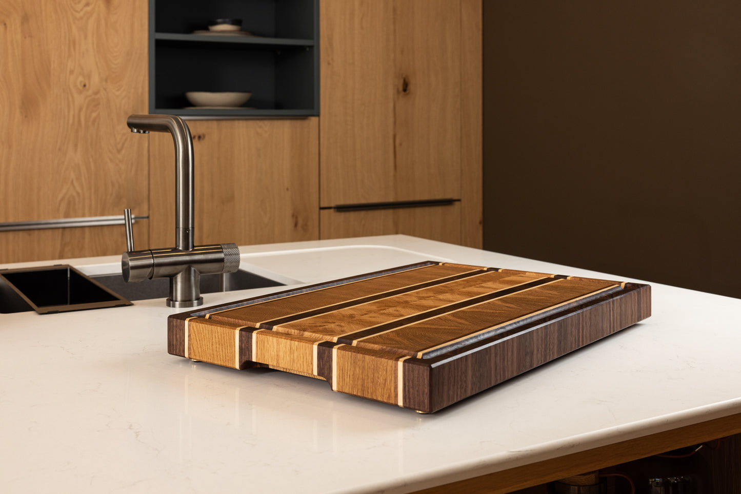 Vue latérale de la planche à découper Ravel en chêne, noyer et érable. Le design graphique du bois de bout et les finitions soignées soulignent son caractère artisanal et luxueux. Fabriquée en France, posée sur un plan de travail de cuisine moderne