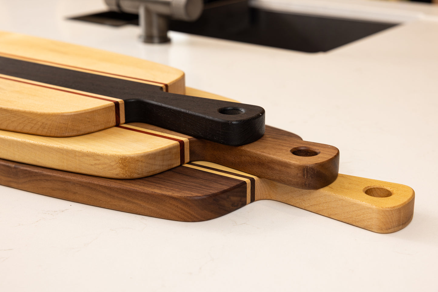 Ensemble de planches apéro artisanales en bois massif, empilées sur un plan de travail, avec double poignées et design graphique en multi-essences.
