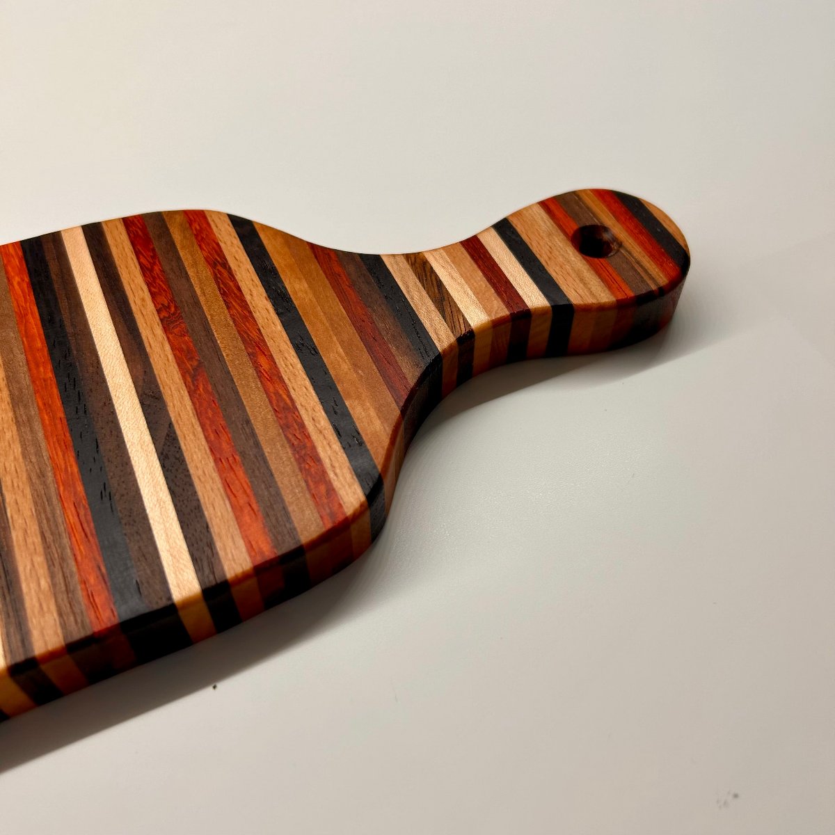 Détail de la poignée d’une mini planche artisanale haut de gamme en bois, design à rayures multi-essences, parfaite pour la cuisine et les apéritifs.