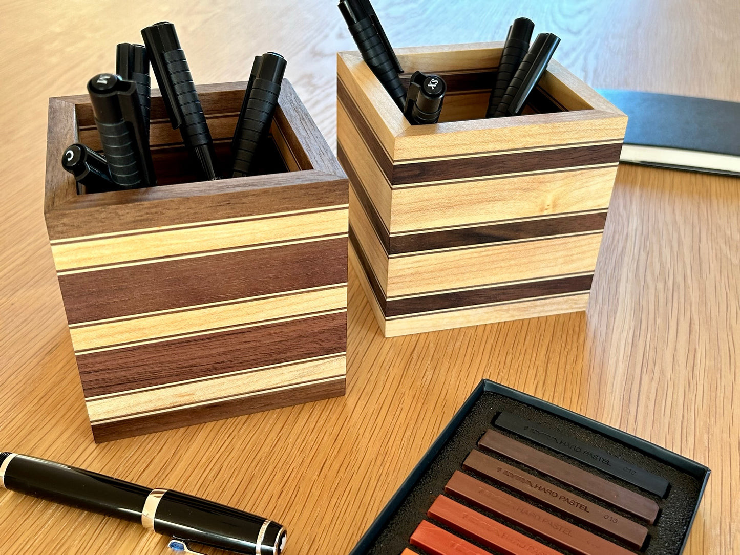 Vue des deux variantes du pot à crayons Debussy, en noyer et érable, présentées côte à côte avec des stylos plume et des feutres