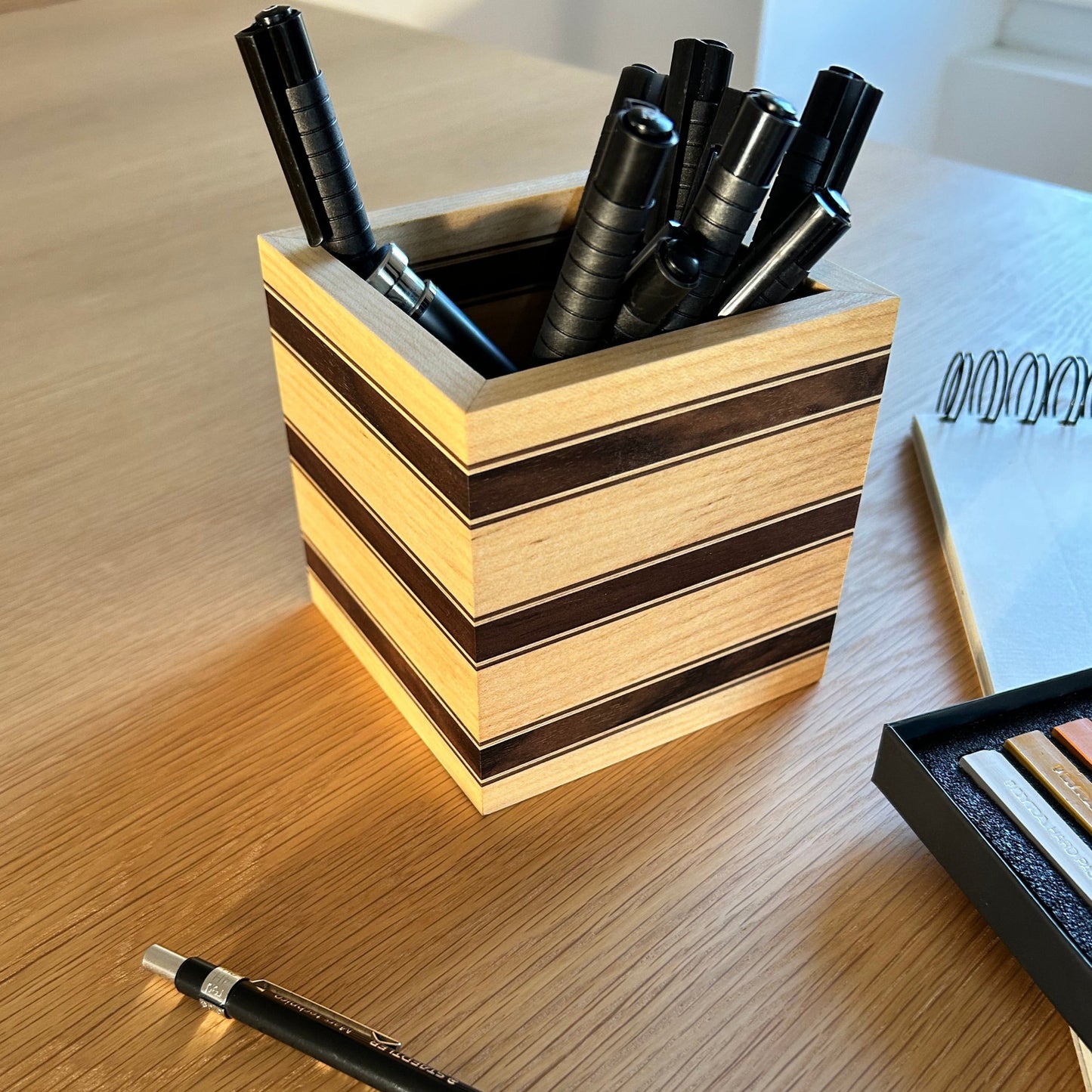 Pot à crayons Debussy en érable avec des bandes fines en noyer américain, en situation avec des stylos et des articles de papeterie