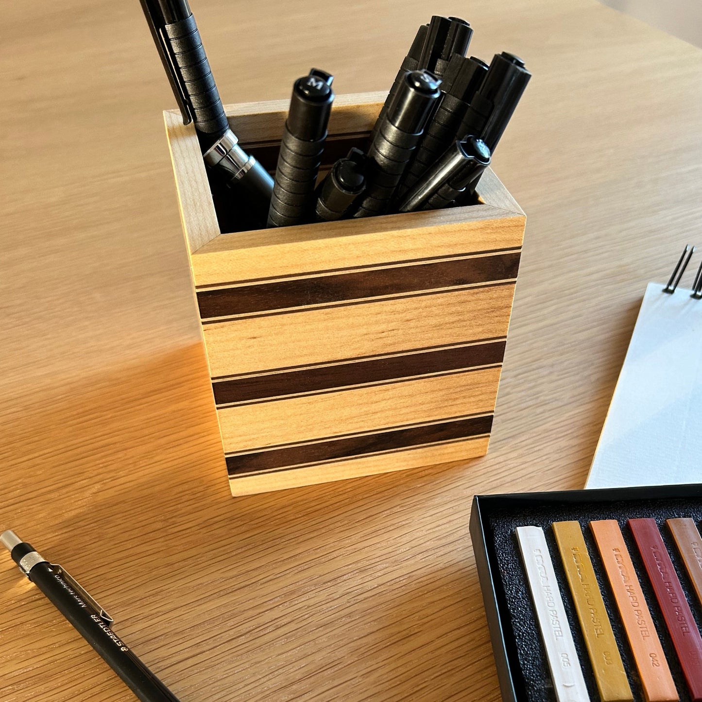 Vue rapprochée du pot à crayons Debussy en érable avec des bandes fines en noyer américain, contenant des stylos et des pastels aux tons bruns