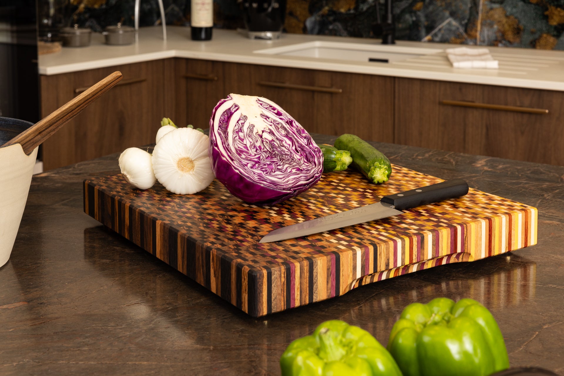 Planche artisanale de luxe en bois de bout 11 essences, en situation sur un plan de travail de cuisine en granit, avec légumes et couteau de chef.