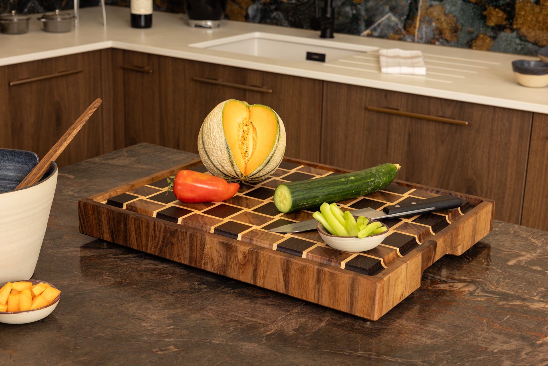 Planche à découper en bois de bout modèle Berlioz, en wengé, noyer et érable, posée sur un plan de travail en granit. Mise en scène avec légumes frais et couteau de chef, pièce artisanale française en bois haut de gamme
