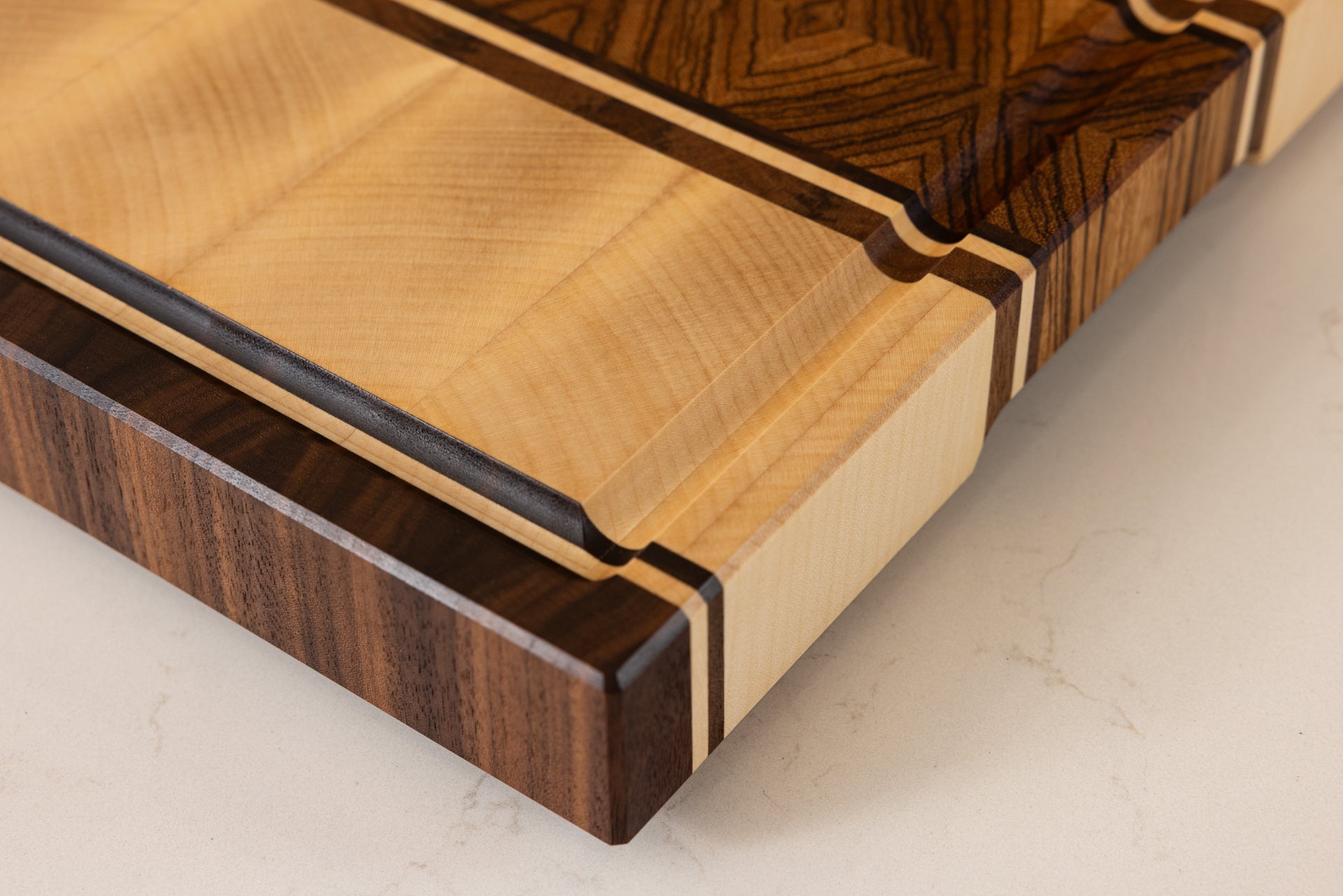 Détail d’une planche à découper artisanale en bois massif, modèle Couperin, montrant le coin avec rigole à jus sculptée, en zebrano, érable et noyer américain, posée sur un plan de travail de cuisine
