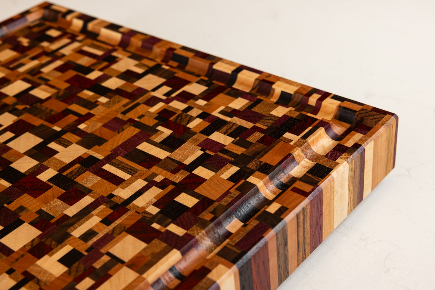 Détail premium de la planche à découper Messiaen en 9 essences de bois précieux et colorés, vue du coin avec rigole à jus fonctionnelle et finitions des bords biseautés élégamment.