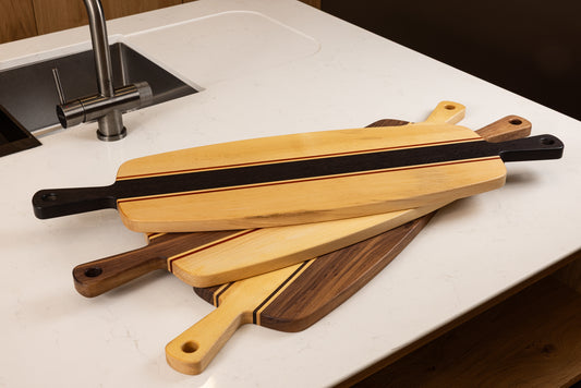 Ensemble de planches apéro artisanales en bois massif, aux finitions graphiques contrastées et doubles poignées, posées sur un plan de travail en cuisine.
